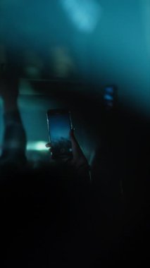 Canlı müzik konseri kaydeden, akıllı telefon tutan bir hayranın fotoğrafını çek. Kalabalık arka planda, elleri havada, dramatik mavi sahne ışıkları altında görülüyor. Dikey video.