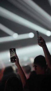 Canlı müzik konserindeki kalabalığın dikey çekimi. Taraftarlar akıllı telefonları bekletip gösteriyi kaydediyorlar. Siluetler parlak, yanıp sönen sahne ışıkları ve atmosferik dumana karşı görünür..