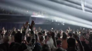 Büyük, enerjik bir kalabalığın video klibi bir müzik festivalinde parlak sahne ışıklarının karşısında. Hayranlar dans ediyor, alkışlıyor ve telefonlarla kayıt yapıyorlar. Etkileyici, yüksek enerjili atmosfer.