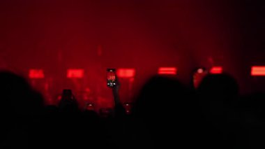 Canlı müzik konserindeki siluetli kalabalık programı kaydetmek için akıllı telefonları soyuyor. Sahne yoğun, yanıp sönen kırmızı ve beyaz flaş ışıklarıyla aydınlanıyor ve enerjik bir atmosfer yaratıyor..