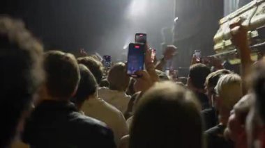 Canlı müzik konseri veya festivalinde heyecanlı bir kalabalığın arka görüntüsü. Birçok taraftar akıllı telefonlarını kaldırarak sahnedeki performansını kaydediyor ve karanlık mekanda parlayan ekranlardan oluşan bir deniz yaratıyor..