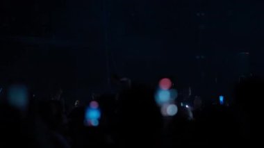 Canlı müzik konserindeki siluetli kalabalık. Hayranlar dans ediyor, el sallıyor ve sahneden parlak mavi ve kırmızı flaşlı ışıklar gibi telefonlarla kayıt yapıyor ve enerjik bir parti atmosferi yaratıyor..