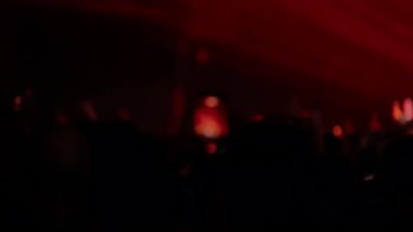 Video, kırmızı sahne ışıkları olan bir konserde bir grup silueti gösteriyor. Bir taraftar programı kaydetmek için bir telefon tutuyor, odak ekranın geniş açısından yakın açısına kayıyor.