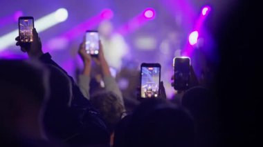Canlı müzik konserindeki kalabalığın görüntüsü. Taraftarlar programı kaydetmek için akıllı telefonları kaldırıyorlar, sonra da tezahürat ve dans için ellerini kaldırıyorlar. Siluetler parlak mor ve mavi sahne ışıklarına karşı.