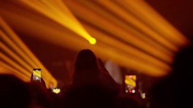 Canlı müzik konserindeki kalabalığın arkasından bir video. Taraftarlar gösteriyi kaydetmek için telefonları tutuyor. Sahne ışıkları yanıp sönen, göz kamaştırıcı bir beyaz ışıkla son buluyor. Enerjik atmosfer.