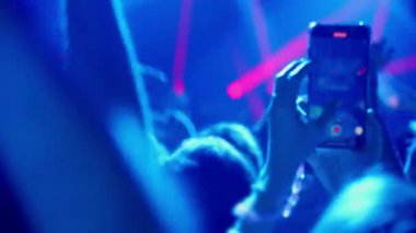 Canlı müzik konseri ya da festivaldeki bir kalabalığın videosu. İnsanlar dans eder, ellerini kaldırır ve canlı sahne ışıkları farklı renklerde yanıp sönerken akıllı telefonlarıyla şovu kaydederler..