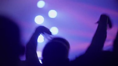 Bir konserde ya da müzik festivalinde dans eden siluetli bir kalabalığın yumuşak odak görüntüleri. İnsanlar dinamik mor ve mavi sahne ışıkları arka planda atmosferik bokeh oluştururken ellerini kaldırıyorlar..
