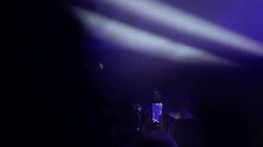 Bir müzik festivali veya konserde siluetli bir kalabalığın görüntüsü. İnsanlar ellerini kaldırır ve performanslarını akıllı telefonlarıyla filme alır. Sahne karanlık.,