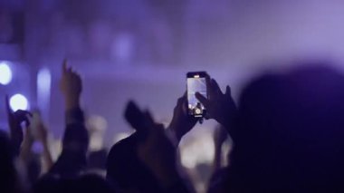 Konserdeki siluetli kalabalık programı akıllı telefonlardan çekiyor. El kamerasıyla çekilen görüntüler, hüzünlü mavi ve mor sahne ışıklarıyla yıkanmış bir müzik performansı kaydeden hayranları yakalıyor..