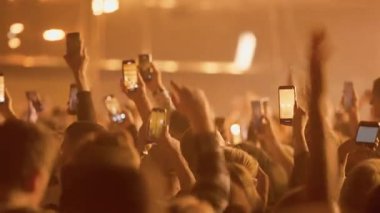 Canlı müzik festivali ya da gösterisindeki büyük bir seyircinin video görüntüleri. Taraftarlar performansı kaydetmek için cep telefonlarını kaldırırken, ekranlar dinamik sahne ışıklandırmasıyla karanlıkta, puslu mekanda parlıyor..