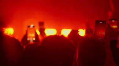 Video sahnesi bir müzik konserinde veya festivalde hayranları gösteriyor. İnsanlar performansı kaydetmek için kırmızı, beyaz ve mavi sahne ışıklarıyla el çırpıyor ve akıllı telefonları tutuyor..