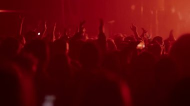 Bir konser veya festivaldeki siluetli kalabalığın videosu. Taraftarlar ellerini kaldırıp akıllı telefonlarla kayıt yaparken sahne yoğun kırmızı ışıktan parlak beyaz arka plana kayıyor..