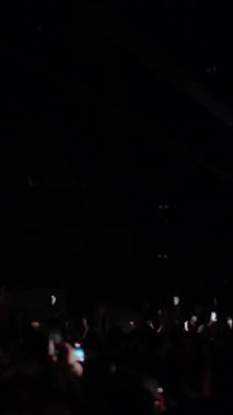 Elleri ve telefonları havada bir konser kalabalığının el kamerasıyla çekilmiş görüntüsü. Dramatik sahne ışıkları yanıp sönerken, yukarıda büyük bir hoparlör dizisi asılı. Enerjik ve atmosferik canlı bir müzik yaratıyor.