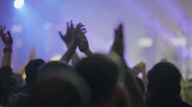 Video sahnesi, bir konserin keyfini çıkaran siluetli bir kalabalığı gösteriyor. Sahne, beyaz ışıklarla karanlıktan canlı ve renkli bir ışık gösterisine dönüşüyor. Elleri havada, bir festivalin enerjisini yakalıyor..