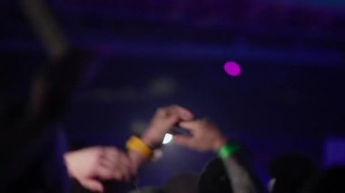 Canlı bir etkinlikteki kalabalığın yumuşak odaklı görüntüleri. Hayranlar mavi sahne ışıkları altında el çırpıp el sallıyorlar. Bir telefon feneri olay yerini aydınlatıyor. Enerjik, duygusal ve atmosferik titreşim.