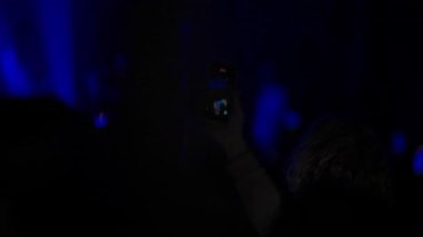 Karanlık bir konser topluluğundaki biri programı çekmek için akıllı telefon kaldırır. Sahne canlı mavi ışıkla yıkanmış ve telefon ekranında canlı performansın kaydedildiği görülüyor. Gece hayatı görüntüleri..