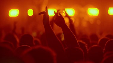 Video görüntüleri, bir müzik festivalinde elleri havada bir grup silueti gösteriyor. İnsanlar canlı turuncu ve sarı sahne ışıkları altında dans ediyor ve tezahürat yapıyorlar.,