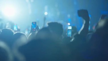 Canlı müzik programındaki siluetli izleyiciler akıllı telefonlarındaki performansı filme alıyor. Parlak, puslu ışıktan, mavi sahne ışıklarıyla karanlık, atmosferik bir sahneye geçiş..