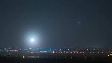 Bir yolcu uçağı gece aydınlık bir havaalanı pistine iniyor ve güçlü iniş ışıkları karanlığı aydınlatıyor.