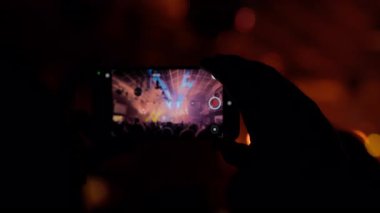 Bir insan silueti akıllı bir telefonla dinamik bir konser kaydeder, canlı sahne ışıklarını yakalar, mavi, kırmızı ve enerjik kalabalık atmosferini yakalar.