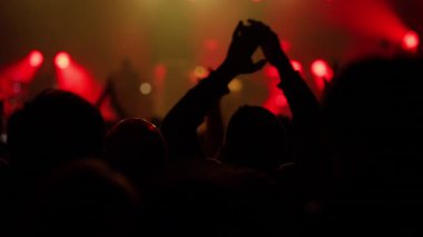 Canlı canlı bir müzik konserinde siluetli kalabalık. El çırpıyor ve heyecanla yükseliyor. Dinamik kırmızı ve sarı sahne ışıkları arka plandaki sanatçıları aydınlatır