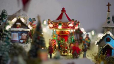 Çekici, aydınlık, minyatür bir Noel köyünün yakın çekim videosu. Merkezinde karla kaplı evler ve bir kilise ile çevrili kırmızı ve beyaz renkli bir atlıkarınca dönüyor.