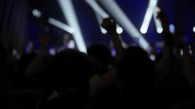 Canlı konserdeki enerjik kalabalık, pek çok kişi gösteriyi çekmek ya da fotoğraflamak için akıllı telefonları tutuyor. Dinamik mavi ve beyaz sahne ışıkları karanlık mekanı aydınlatarak heyecan verici bir atmosfer yaratıyor.