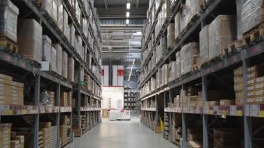 Kamera büyük, modern bir depo koridorunda hareket ediyor. Uzun metal raflar karton kutular ve envanter ile dolu, lojistik, depolama ve tedarik zinciri yönetimi sergileniyor. Kimse yok..