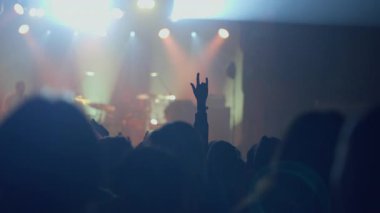 Siluetli konsere gidenler el kaldırır, bazıları parlayan ışıklarla aydınlatılmış bir sahneye karşı rock hareketleriyle el kaldırır. Canlı bir müzik etkinliğinin enerjik atmosferini ve keyfini yakalar.