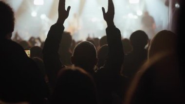 Coşkulu bir seyircinin canlı bir müzik konserinde ellerini başının üstünde çırpışının arka görüntüsü. Parlak sahne ışıkları altında canlı performansın keyfini çıkaran insanların siluetleri.