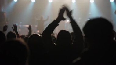 Dinamik bir video canlı müzik konserinde heyecanlı bir seyirci yakalıyor. Parlak bir sahne önünde el çırpma ve sallanma siluetleri enerjik bir atmosfer yaratır.