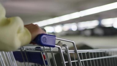 Alışveriş arabasının sapına uzanmış bir kadının yakın çekimi. Parlak bir yeraltı otoparkına doğru itiyor. Sığ alan derinliği arkaplanı bulanıklaştırır
