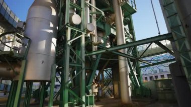 Büyük bir endüstriyel işleme tesisinde düşük açılı çekim. Açık mavi gökyüzüne karşı karmaşık boru, tank ve çelik yapılar ağı. Üretimi, enerjiyi ve mühendisliği temsil eder.