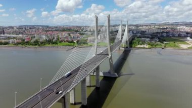 Portekiz, Lizbon 'daki Vasco da Gama Köprüsü' nü gösteren hava aracı videosu geri çekiliyor. Çekimde Tagus Nehri üzerinde modern mimari sergileniyor. Şehir mavi gökyüzünün altında ufuk çizgisi bulunuyor.
