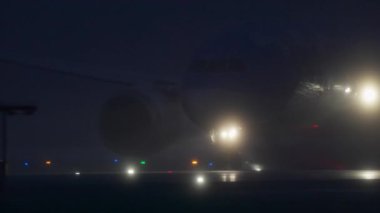 Islak bir pistte, gece yoğun siste yavaşça ilerleyen ticari bir uçak görülüyor. Parlak iniş ışıkları ve yer ışıkları yolu aydınlatıyor ve etrafındaki sisi aydınlatıyor.