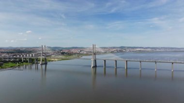 Portekiz, Lizbon 'daki ikonik Vasco da Gama Köprüsü' ne doğru uçan hava aracı görüntüleri. Kamera, güneşli bir günde Tagus Nehri üzerindeki modern mimariyi yakalar.