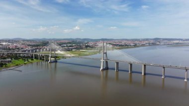Portekiz, Lizbon 'daki Tagus Nehri üzerindeki görkemli Vasco da Gama Köprüsü' ne doğru uçan hava aracı görüntüleri. Modern mimariyi, şehir siluetini ve güneşli bir günde manzarayı gösterir