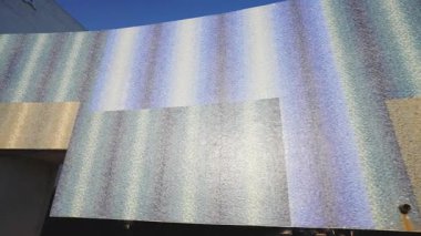 Parıldayan, yanardöner mozaik fayanslarla kaplı büyük, eğimli bir binanın görüntüsü. Kiremitlerin açık bir gökyüzünün altında mavi, beyaz ve altından oluşan dalgalı bir deseni var.