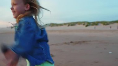 Güzel bir günbatımında kumlu bir plajda özgürce koşan sarı saçlı neşeli bir genç kız. Bu çekim onun kaygısız ruhunu, enerjisini ve çocukluğun saf mutluluğunu yakalar.