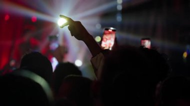 Canlı bir konserde siluetli bir kalabalığın yakın çekimi, pek çok kişi kaydetmek için akıllı telefonları tutuyor. Kalkmış bir el, yavaş çekimde çekilen parlak sahne ışıklarıyla aydınlatılır.