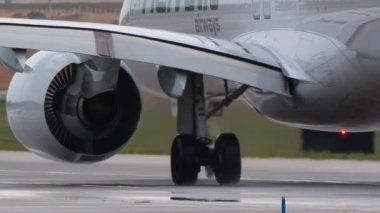 Islak bir pistte taksicilik yapan geniş gövdeli bir yolcu uçağının yakın plan telefon çekimi. Motorun, kanadın ve iniş takımlarının detaylı görüntüsü. Tekerleklerden su püskürtülmüş. Isı sisi havada parıldıyor..