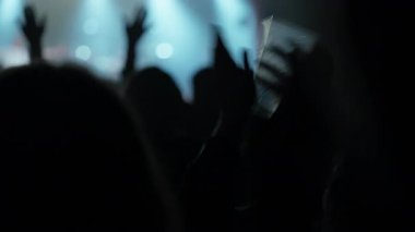 Siluetli kalabalık bir müzik konseri veya kulübünde coşkuyla dans ediyor. Yanıp sönen sahne ışıkları dumanlı atmosferi aydınlatır. Bir kişi plastik bardağı tutar ve müziğe doğru sallanır.