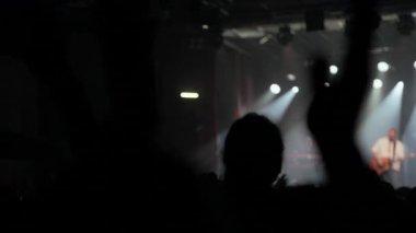 Konsere gidenlerden oluşan enerjik bir kalabalık canlı müzik eşliğinde ellerini kaldırıp alkışlıyor. Hayranların siluetleri, müzisyenler ve dumanlı atmosferli parlak bir sahne karşısında dinamik bir ön plan oluşturuyor.