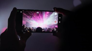 Eller akıllı telefonu tutuyor, pembe ve mor sahne ışıklarıyla canlı bir konser kaydediyor ve tezahürat yapan kalabalığı aydınlatıyor. Bir müzik festivalinde ya da partide mobil bir cihazla enerjik anları yakalamak