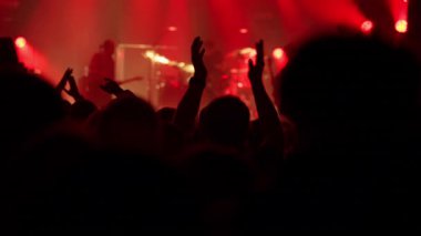 Seyircilerin arkasından çekilen canlı bir konserde heyecanlı bir kalabalık. Siluetli eller kaldırılır ve canlı kırmızı, turuncu ve sarı sahne ışıkları altında ağır çekimde alkışlanır.