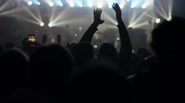 Canlı konser ya da müzik festivalinde siluetli bir kalabalığın dinamik çekimi coşkuyla tezahürat yapıyor, alkışlıyor ve heyecanla el kaldırıyor. Parlak sahne ışıkları puslu arkaplanı aydınlatır