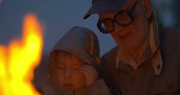 Oudere man zit met kleine jongen in de buurt van kampvuur — Stockvideo