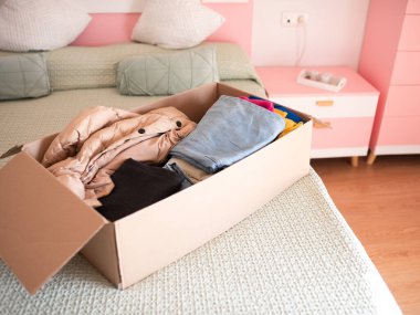 Karton kutuda ceketler ve pantolonlar da dahil olmak üzere çeşitli giysi parçaları var. Pastel desenli ve yumuşak dokuları olan rahat bir yatak odasında düzgünce yapılmış bir yatak odası.