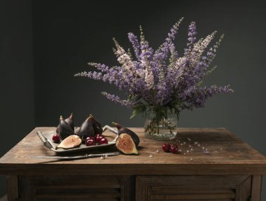 Olgun incirlerle ve vazoda kır çiçekleriyle nefis bir kompozisyon klasik resim tarzı karanlık bir tabloda eski ahşap masa üzerinde..