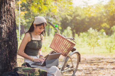 Yeşil atletli güzel Asyalı kadını kapatın. Halka açık bir parkta dizüstü bilgisayarla ağacın altında beyaz bir bisikletin yanında çalışıyor.